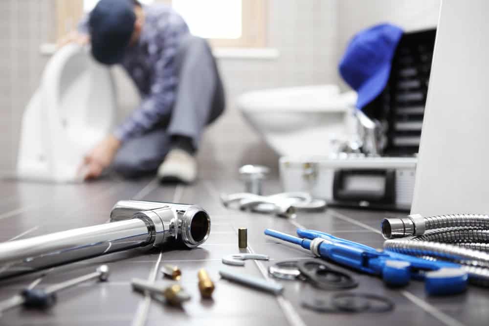 A plumber works on installing a toilet in a bathroom. In the foreground, various tools and plumbing components, including pipes, wrenches, and screws, are spread out on the tiled floor. A toolbox and a blue cap are visible near the plumber specializing in drain cleaning IL.