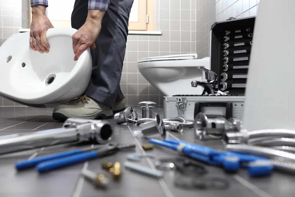 A person in a bathroom is installing a white sink. Various plumbing tools and parts, including wrenches, pipes, and hose fittings, are scattered on the tile floor. A tool kit is open beside a toilet in the background as the Plumber IL works diligently on this detailed installation.