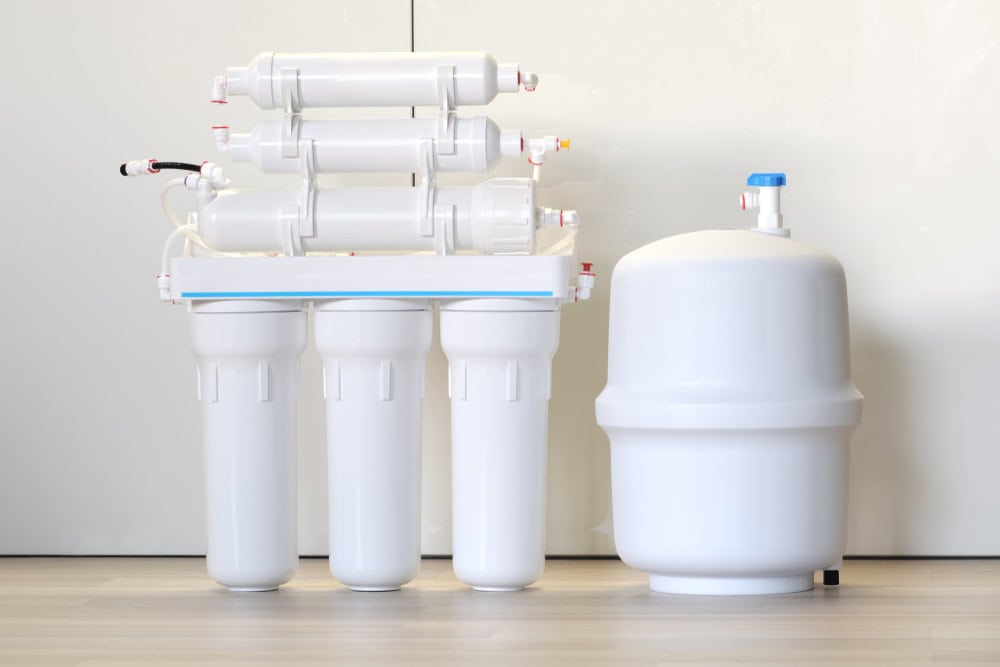 A reverse osmosis water filtration system with four vertical cylindrical filters connected to a series of horizontal filters on top, alongside a white storage tank with a blue valve, all placed on a light-colored floor against a plain wall background, flawlessly installed by an expert plumber in IL.