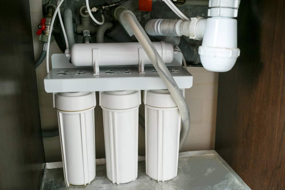 The image shows an under-sink water filtration system with multiple cylindrical filter cartridges connected by various tubes and valves. It is installed beneath a kitchen sink, which also appears to have had a recent Drain Cleaning IL, with some plumbing pipes visible on the right side.