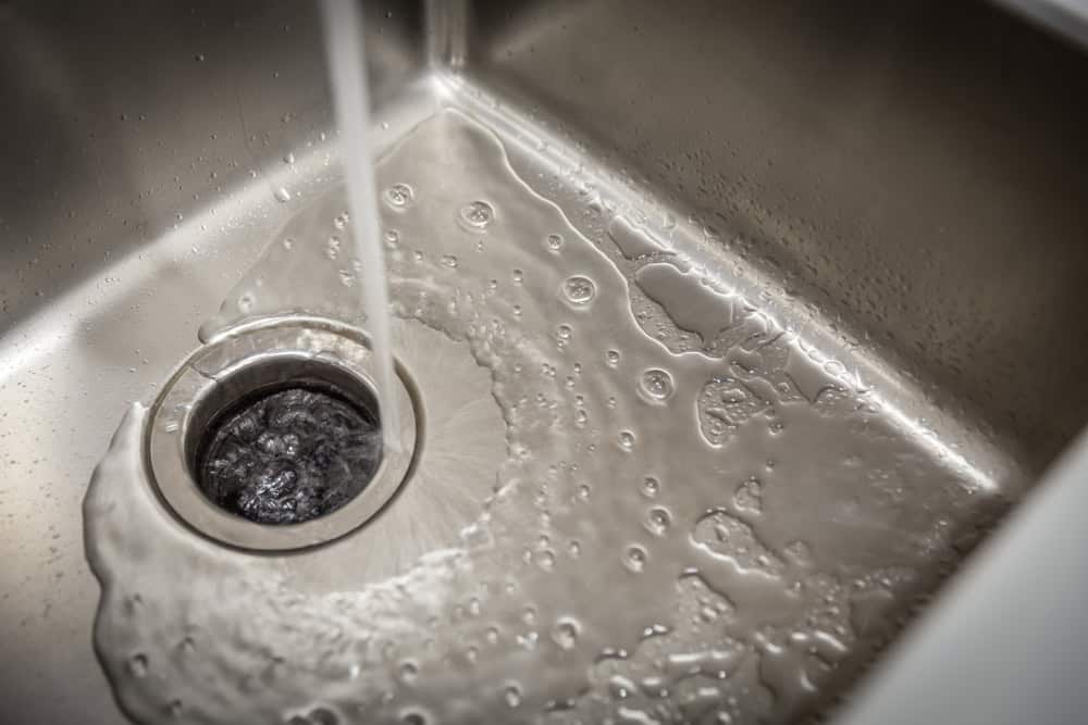 Close-up of a stainless steel sink with water running from the tap into the drain, creating ripples and bubbles around the drain opening. The sink appears clean and well-lit, showcasing the professional touch often seen in Plumber IL services.