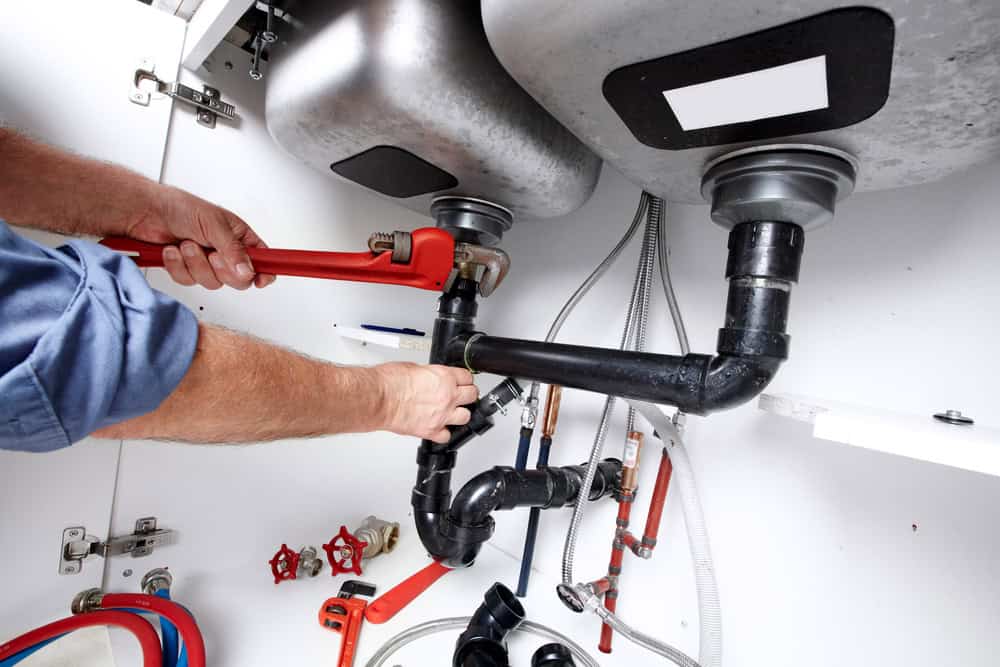 A plumber in IL uses a wrench to tighten pipes under a kitchen sink. Various tools and pipes are visible, including the sink's underside, valves, and connectors. The plumber's forearms and part of a blue sleeve are shown as they work on the plumbing, showcasing their expertise in Sump Pump Installation IL.