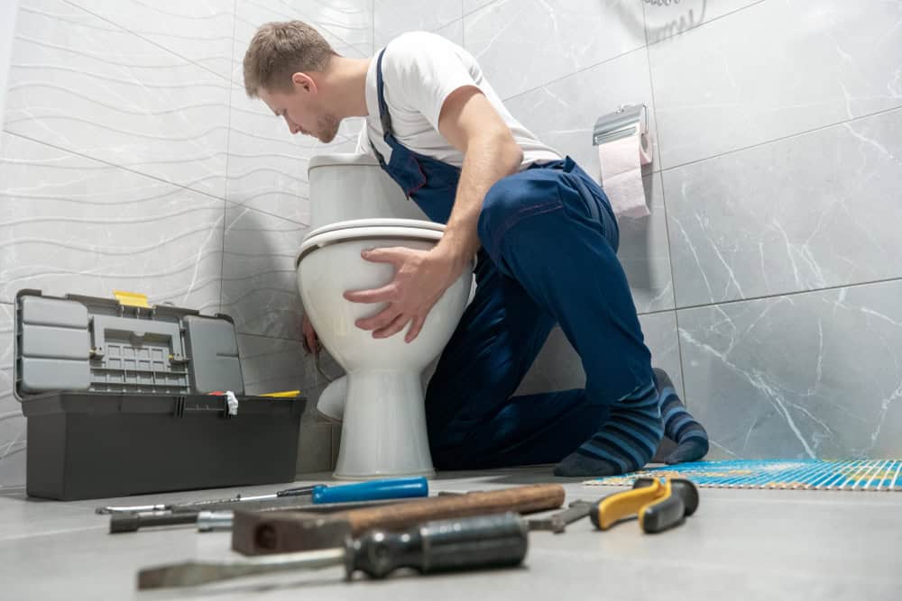 A plumber in blue overalls is kneeling beside a toilet, appearing to repair it. He has a toolbox open on the floor next to him, with various tools spread around, including a wrench, screwdriver, and hammer. The bathroom features gray tiles. This Plumber IL offers expert services like sump pump installation and water heater installation.