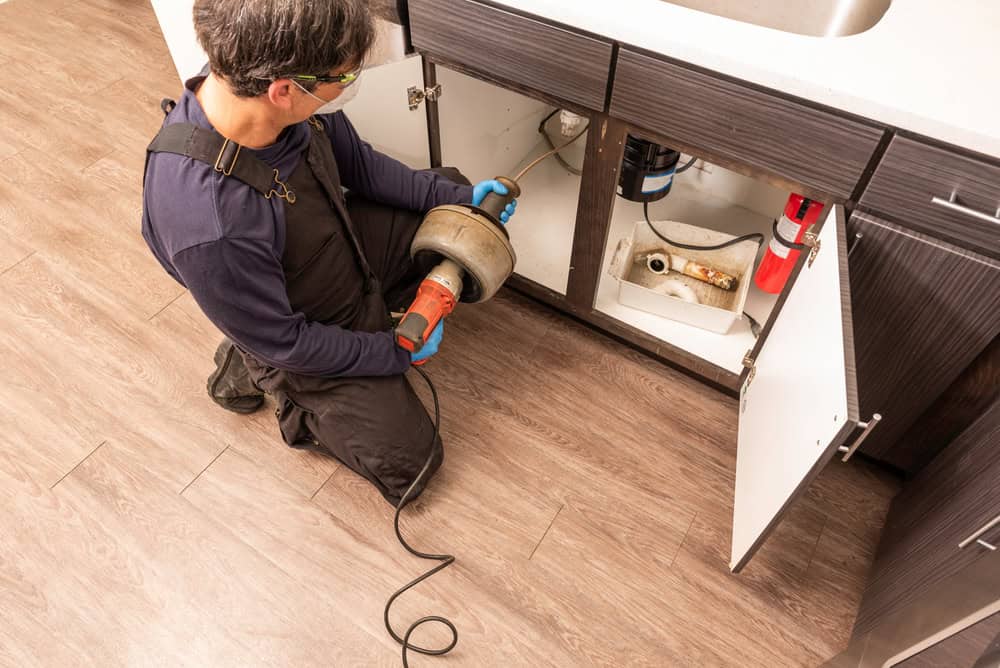 A plumber IL wearing gloves, safety glasses, and overalls uses a drain snake tool to unclog a pipe under a kitchen sink. The cabinet doors are open, exposing the pipes and plumbing beneath as the plumber kneels on a wooden floor.