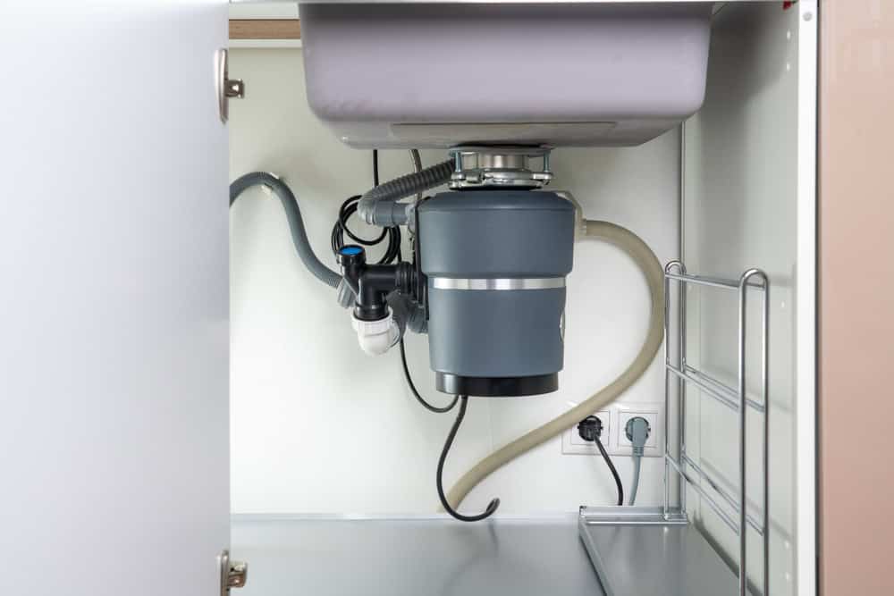 A garbage disposal unit is installed under a kitchen sink. The unit is grey with various black and white pipes connected to it, handled by a professional Plumber IL. There are electrical outlets and a metal rack mounted on the right side of the cabinet. The cabinet door is partially open.