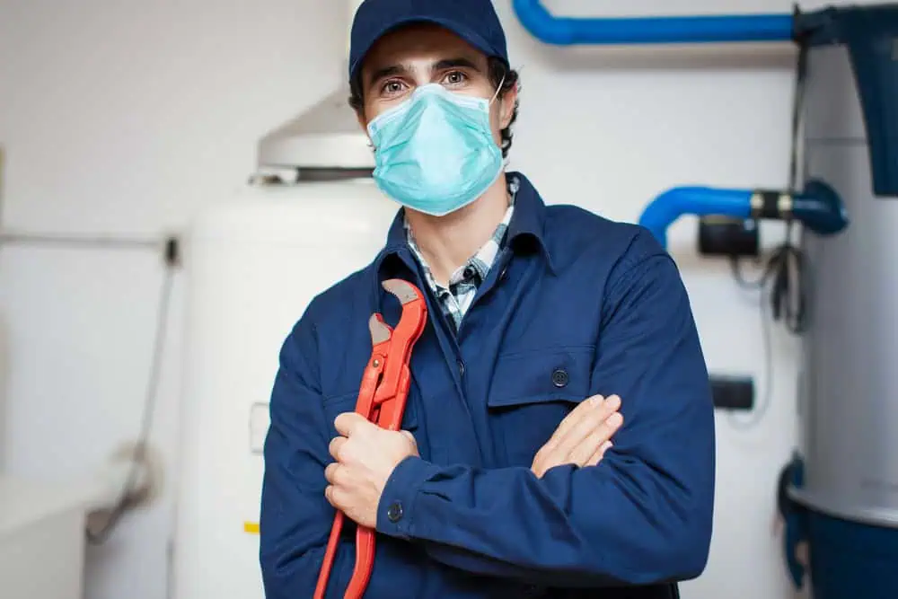 A plumber in blue work overalls, face mask, and cap stands confidently with arms crossed, holding a red pipe wrench. Behind him, plumbing equipment and visible pipes highlight his expertise in services such as sump pump installation, water heater installation, and general plumbing repairs in IL.