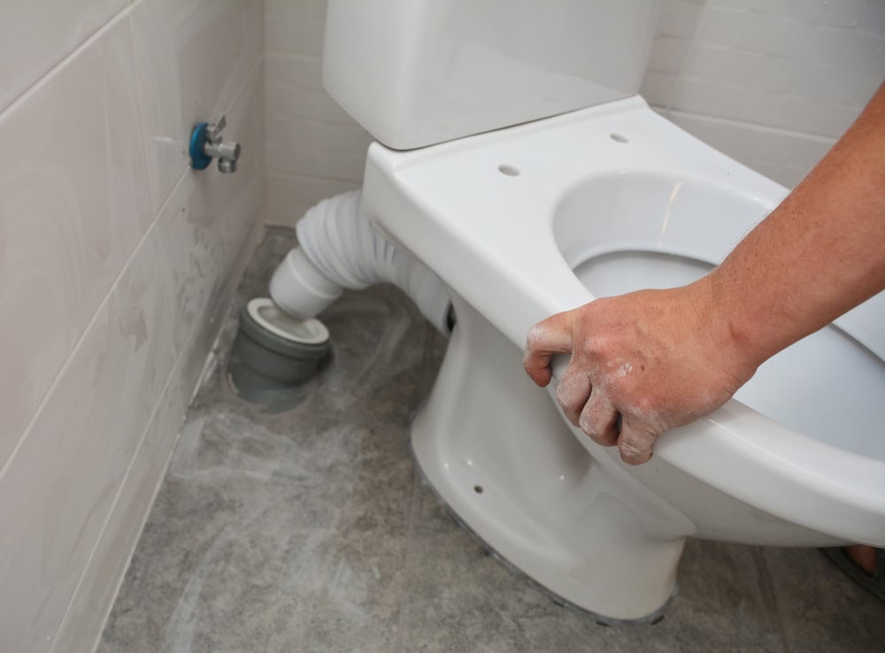 A plumber in IL is installing a white toilet, carefully positioning it over a grey waste pipe connection on a tiled bathroom floor. The person's hands, slightly dusty, grip the base of the toilet bowl to ensure proper alignment.