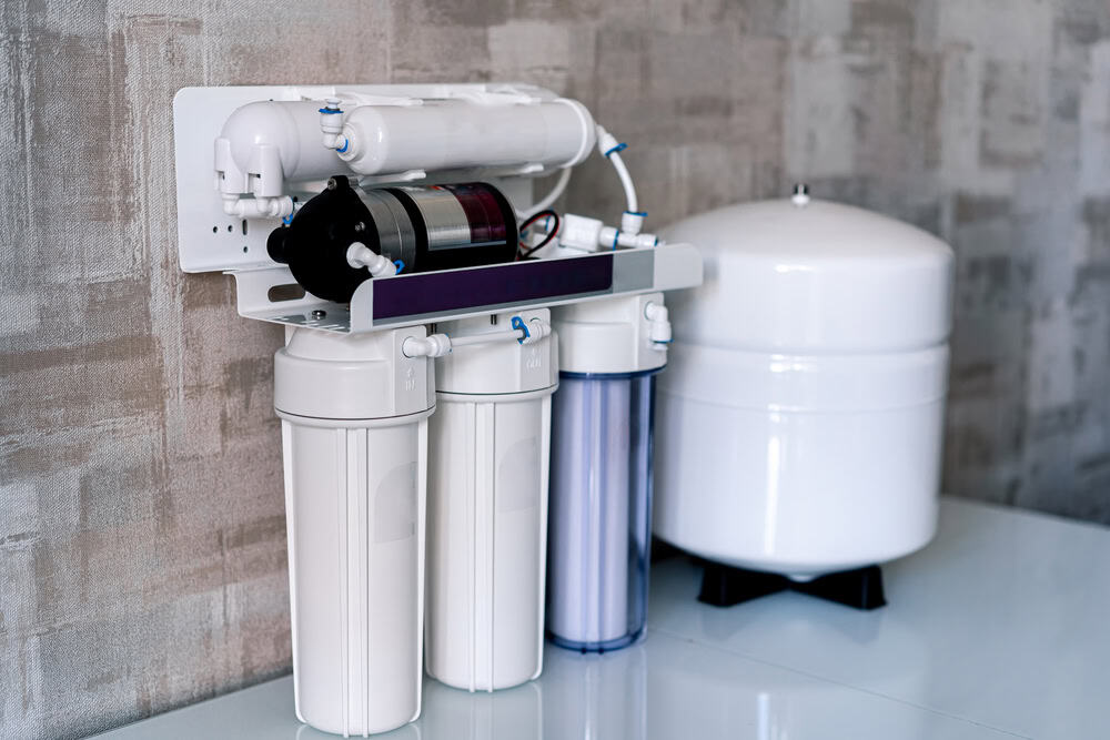 A reverse osmosis water filtration system is mounted on a textured wall, featuring three cylindrical filters and various tubes. A pressurized storage tank sits on the floor next to the system. For related services like Water Heater Installation IL, consult a professional plumber in IL.