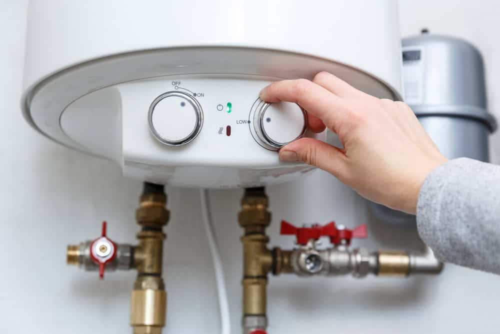 A hand adjusts the temperature knob on a white water heater. The heater has two dials, several brass valves, and pipes beneath it. One valve has a red handle, and a small gray cylindrical component is visible beside the heater—essential for seamless water heater installation in IL.