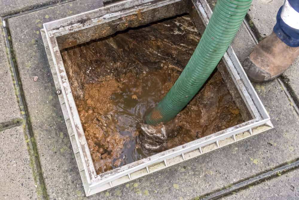 A close-up shot of a person using a green hose to extract waste from a septic tank during a Drain Cleaning IL service. The surrounding ground is made of concrete slabs, and part of the person's shoe is visible on the right edge of the image.