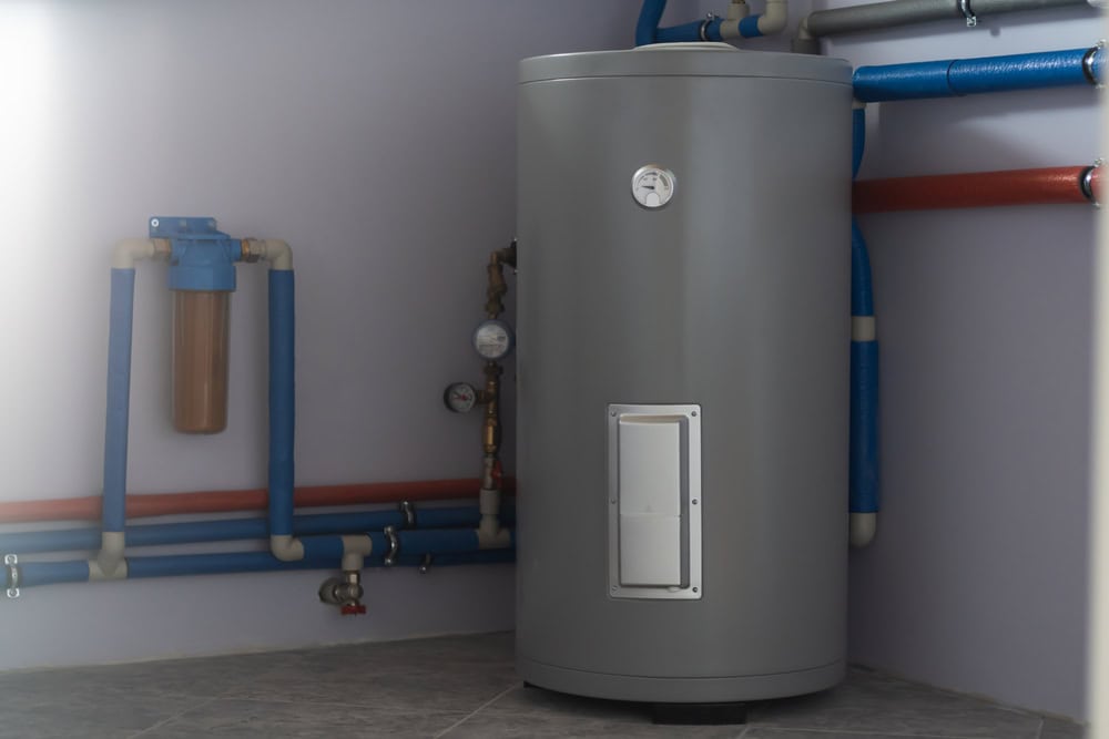 A modern grey cylindrical water heater is situated against a wall with a variety of blue and red pipes connected to it. Visible gauges and valves are attached to the unit and surrounding pipes. The floor is tiled, and the scene is well-lit, showcasing expert workmanship by a Plumber IL.