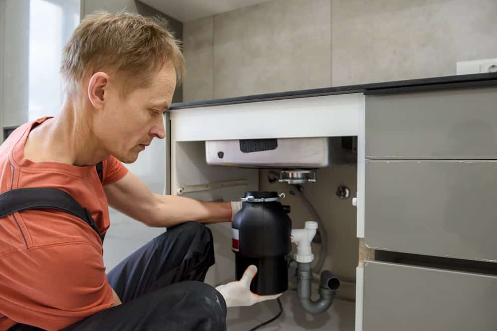 A person with short hair wearing an orange shirt and overalls is installing a garbage disposal unit under a kitchen sink. The cabinet doors beneath the sink are open, revealing various plumbing components, reminiscent of tasks like sump pump installation IL. The countertop and part of the wall are visible.