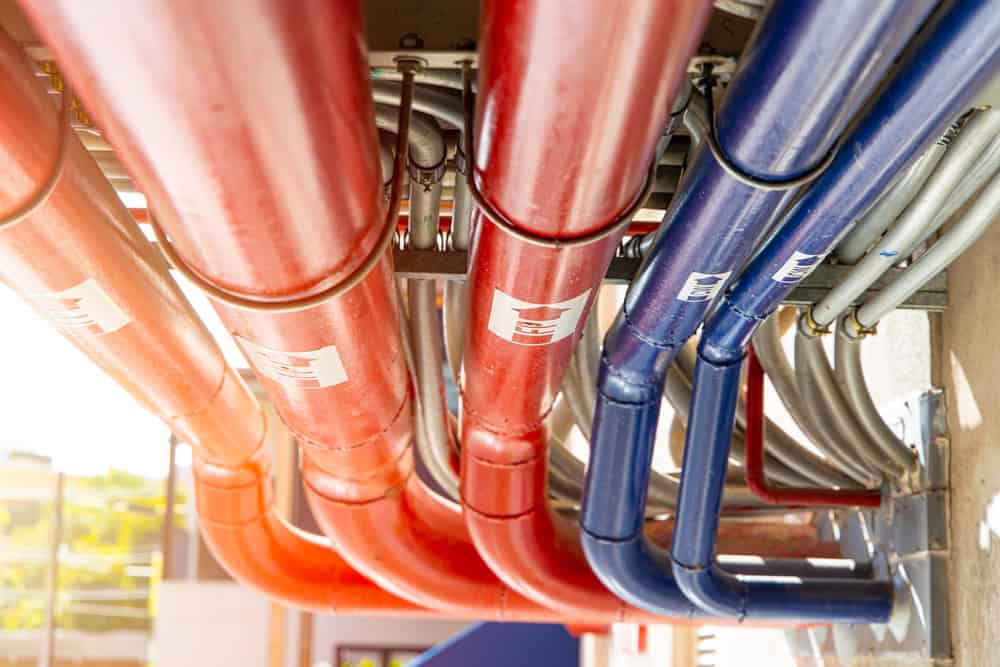 Close-up of red and blue industrial pipes running parallel to each other, fixed on a ceiling, with some labels attached to each pipe. The background is blurred, highlighting the vibrant colors and metallic texture of the pipes, showcasing quality work you’d expect from Plumber IL services.