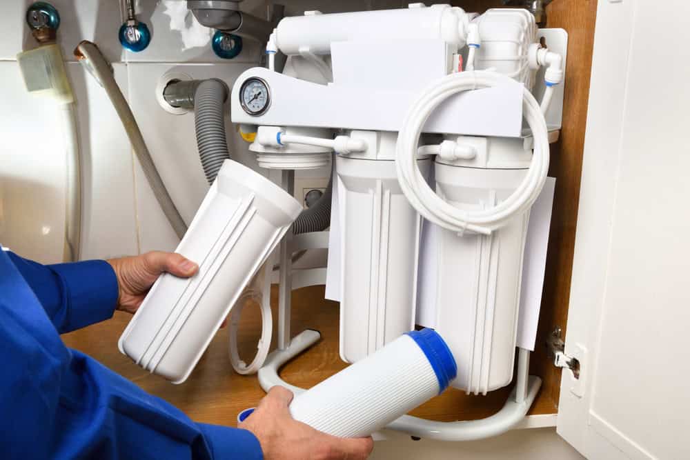 A person in a blue shirt, possibly a Plumber IL, is replacing filter cartridges in an under-sink reverse osmosis water filtration system. Various hoses, pipes, and filtration components are visible beneath the sink. The person holds a used filter in one hand and a new one in the other.