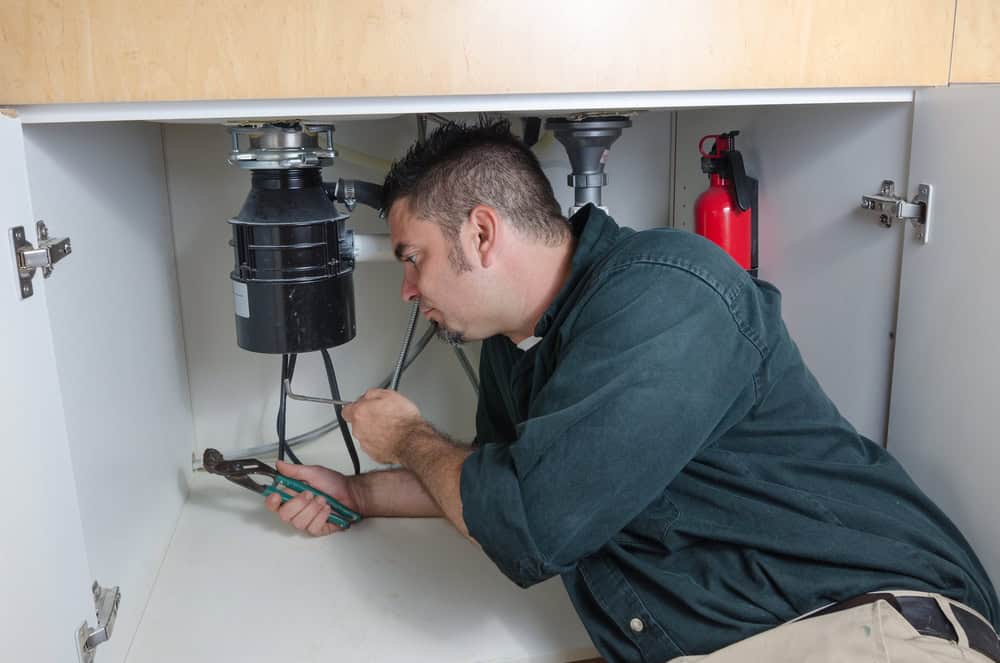 A man is lying on his side under a sink, holding adjustable pliers and working on plumbing pipes connected to a garbage disposal unit. He is wearing a dark green shirt and appears focused on the task, perhaps pondering his earlier Water Heater Installation IL. A red fire extinguisher is visible in the cabinet.