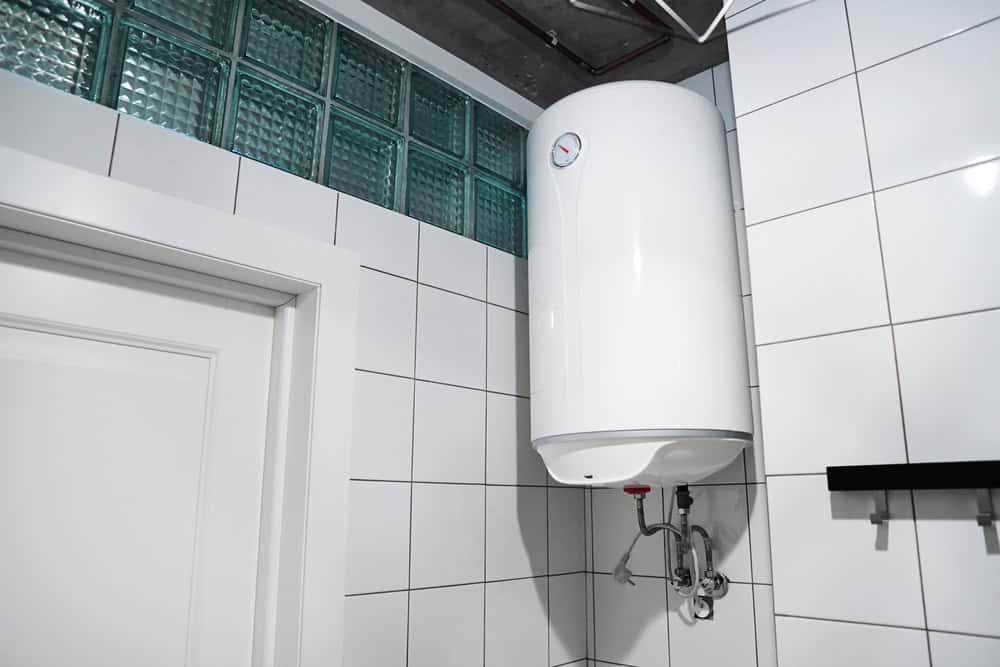 A modern bathroom with white tiled walls features a white cylindrical water heater mounted near the ceiling. The heater has a small gauge for temperature control and is connected by visible pipes. A section of glass block windows can be seen above the heater, installed by a skilled Plumber IL.