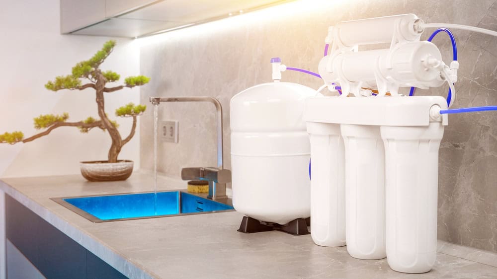 A modern kitchen countertop with a reverse osmosis water filtration system installed beside a sink. The setup includes multiple white filter cartridges and a storage tank. For an added touch, the countertop features a small potted bonsai tree. For professional installation, contact Plumber IL.