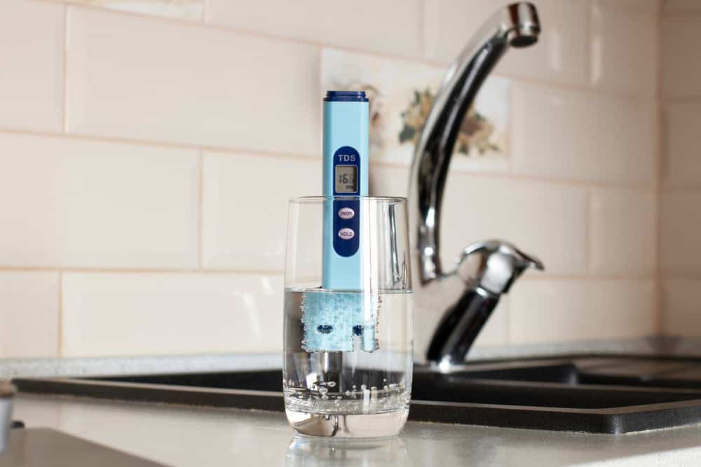 A TDS (Total Dissolved Solids) meter is placed in a glass of water on a kitchen counter, displaying a reading of 15.5 ppm. A sink and faucet are visible in the background, hinting at the need for effective drain cleaning IL.