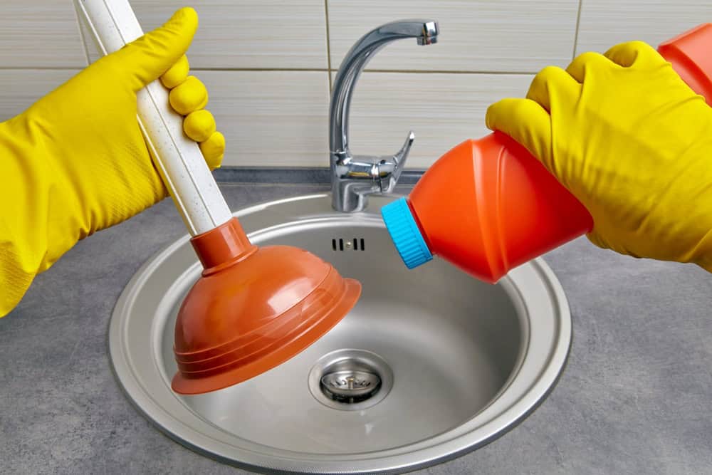 A person wearing yellow rubber gloves holds a red plunger in one hand and a red bottle of drain cleaner in the other, above a stainless steel sink with a modern faucet, perfectly set for mastering Drain Cleaning IL tasks.