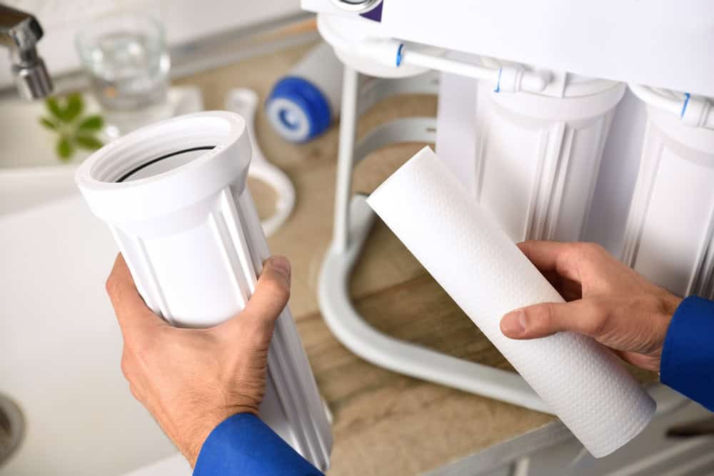 A person replaces the filter cartridge in a water filtration system. The setup includes multiple white filters mounted on a frame. Both hands are involved, one holding the filter housing and the other holding the new cartridge. A kitchen sink is visible in the background, reflecting expert attention akin to Plumber IL services.