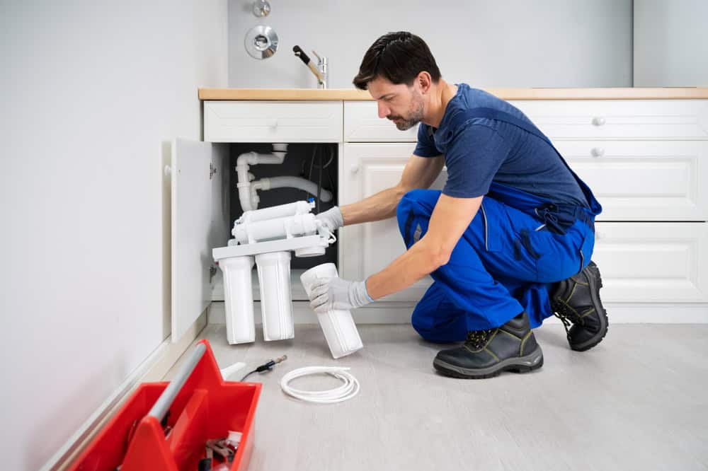 A plumber in IL, wearing a blue uniform and gloves, is kneeling in front of an open cabinet under a sink, installing a water filtration system. Nearby, a red toolbox with various tools is placed on the floor. The kitchen has white cabinets and light-colored flooring.