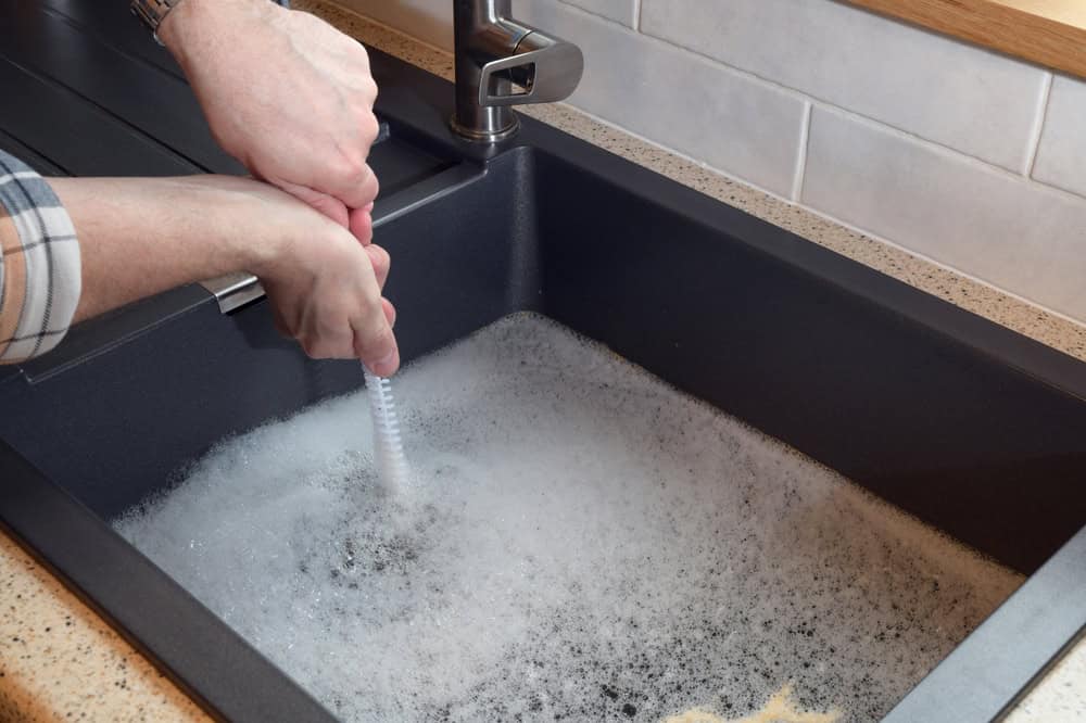 A person wearing a plaid shirt is scrubbing a soapy dish brush in a large black sink filled with sudsy water. The well-lit sink area, featuring a modern faucet and beige countertop with white tiled backsplash, demonstrates the importance of proper drain cleaning in IL homes to keep everything running smoothly.