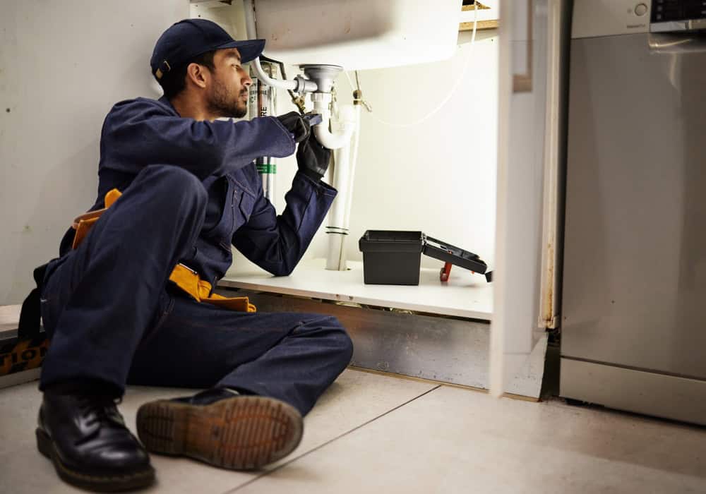 A plumber wearing a cap and work uniform with a tool belt around his waist kneels under a kitchen sink while working on a pipe. Near him is an open toolbox. He is focused intently on his task, exemplifying the same dedication he brings to jobs such as Drain Cleaning IL.