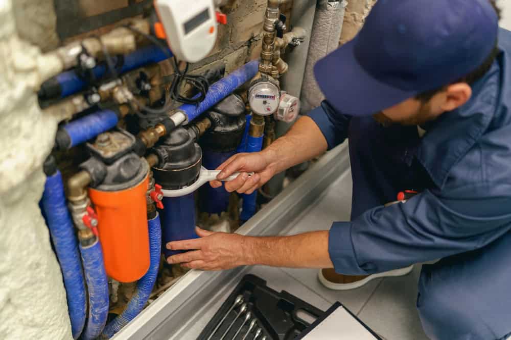 A man in a blue uniform, wearing a cap, is adjusting or repairing a complex array of plumbing pipes and equipment. The setup includes blue and orange filters with various valves and gauges connected by blue hoses—part of a thorough Drain Cleaning IL service.