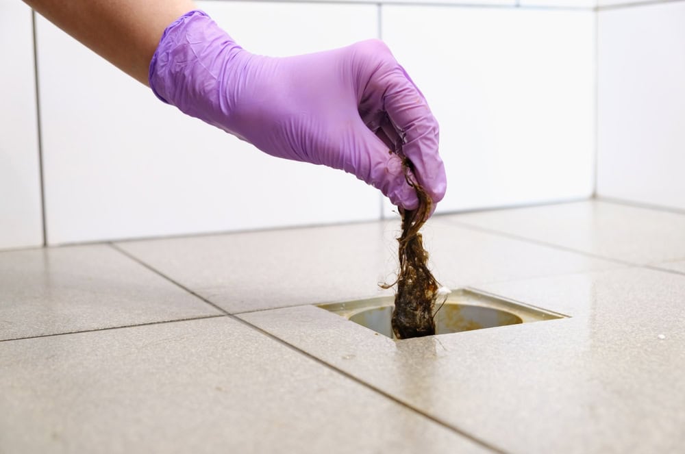 A gloved hand is pulling a clump of hair and debris out of a drain on a tiled floor, showcasing expert Drain Cleaning IL services.