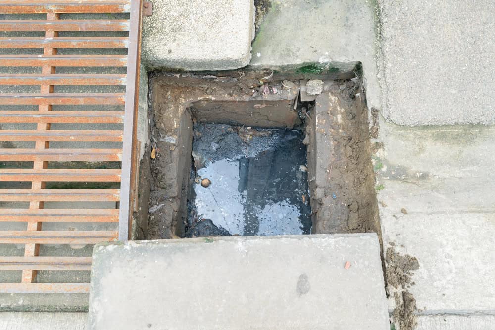 An uncovered rectangular sewage drain filled with murky water is surrounded by concrete and dirt. To the left, there is a rusted metal grate, and to the right, there is a mix of stone and pavement. Debris is visible around the drain, emphasizing the need for professional Drain Cleaning IL services.