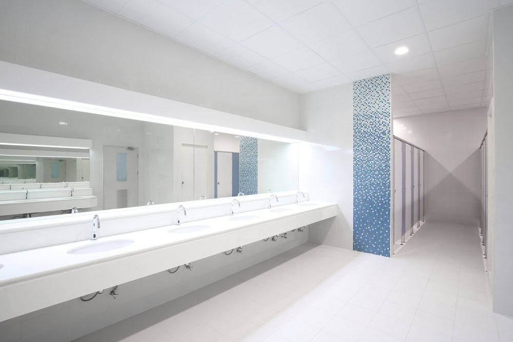 A modern public restroom with a long white counter featuring several sinks and faucets. The wall behind the sinks has a large mirror, and the far side of the room has a row of stalls with blue and white tiled dividers. The room is well-lit with overhead lighting, maintained by Plumber IL for top-notch service.