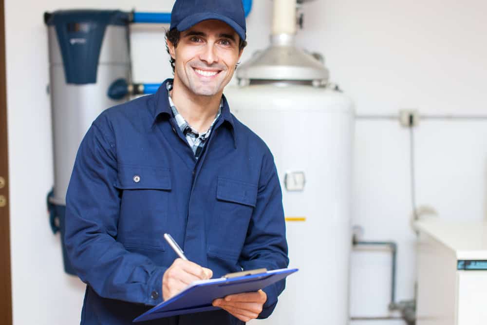 A smiling worker in a blue uniform and cap holds a pen and clipboard. He stands in front of industrial equipment in what appears to be a utility room. The background includes a large water heater, showcasing Water Heater Installation IL services, along with various pipes and fittings.