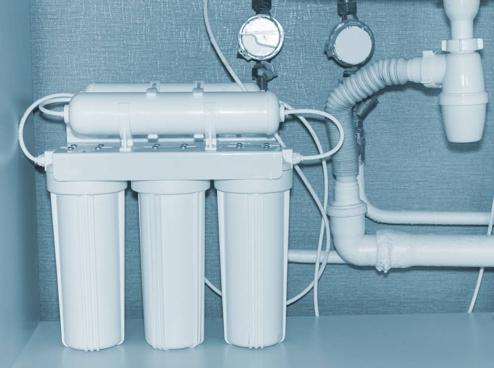 A close-up of a water filtration system installed under a sink. The system includes three cylindrical filter cartridges and connecting pipes. The background shows plumbing, wall-mounted instruments, and an adjacent section for drain cleaning in IL. The setup is clean and neatly arranged.