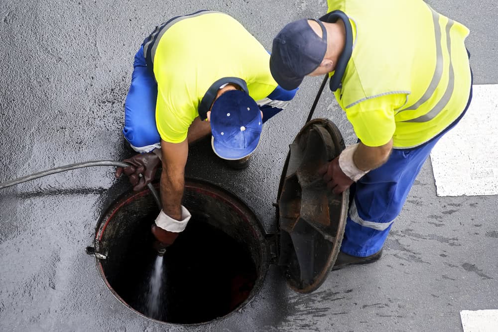 Two workers in high-visibility clothing are seen from above, working on an open manhole. One worker is holding a hose and appears to be spraying inside the manhole, while the other is holding the manhole cover. They seem to be performing thorough drain cleaning IL for optimal maintenance.