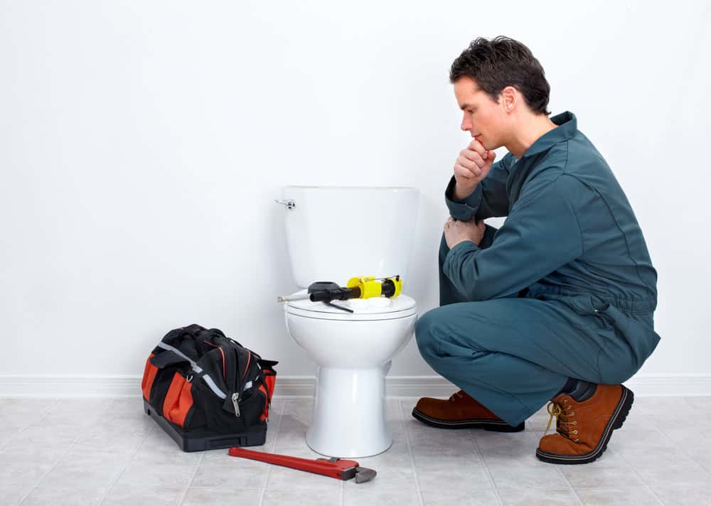 A plumber in a green jumpsuit and brown work boots kneels beside a toilet, thoughtfully inspecting it. On the floor beside him are a toolbox, a pipe wrench, and other plumbing tools, indicative of his expertise in services like Drain Cleaning IL. The background is a simple white wall.