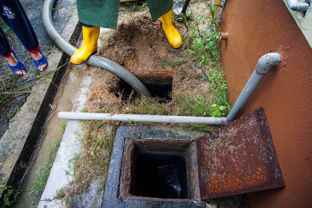 A person wearing yellow rubber boots stands near an open drainage system with a hose inside, possibly conducting a Drain Cleaning IL task. The ground is muddy with grass around the openings, while another person in blue sandals is visible at the edge of the image.