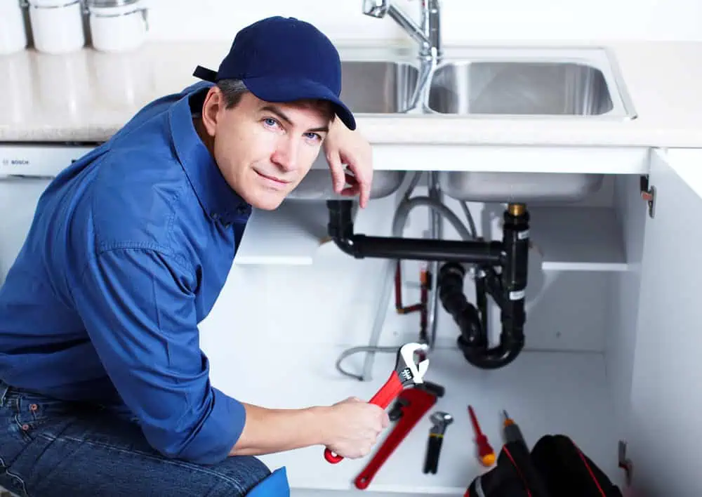 A plumber in a blue uniform and cap holds a wrench while crouching in front of an open cabinet under a kitchen sink. Various tools are scattered around, and the sink pipes are visible.
