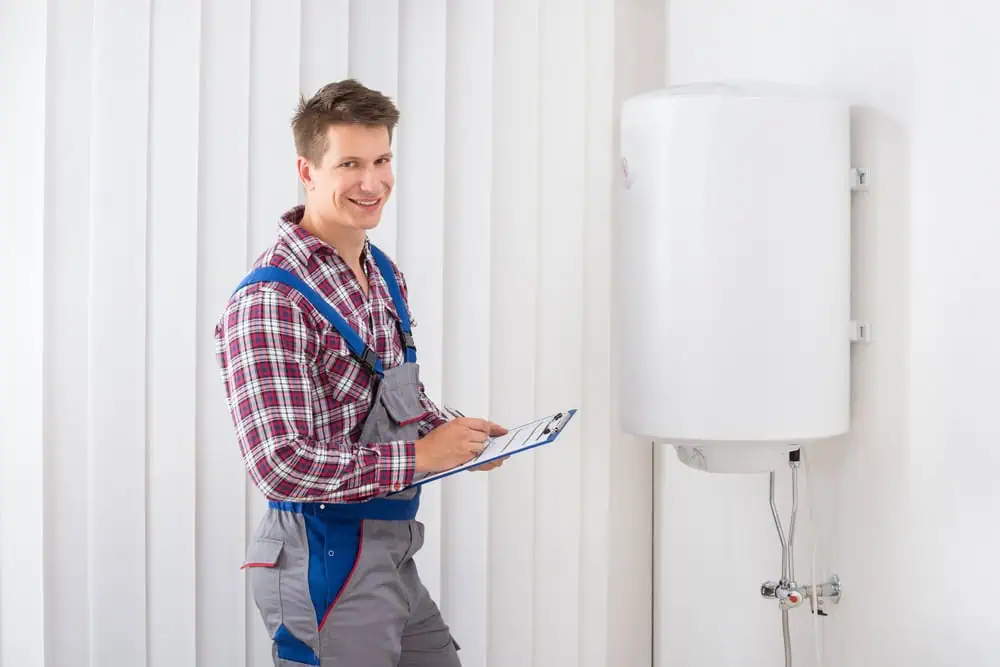 A person in work overalls, wearing a plaid shirt, smiles while holding a clipboard. They stand next to a white boiler mounted on a wall, with vertical blinds in the background.