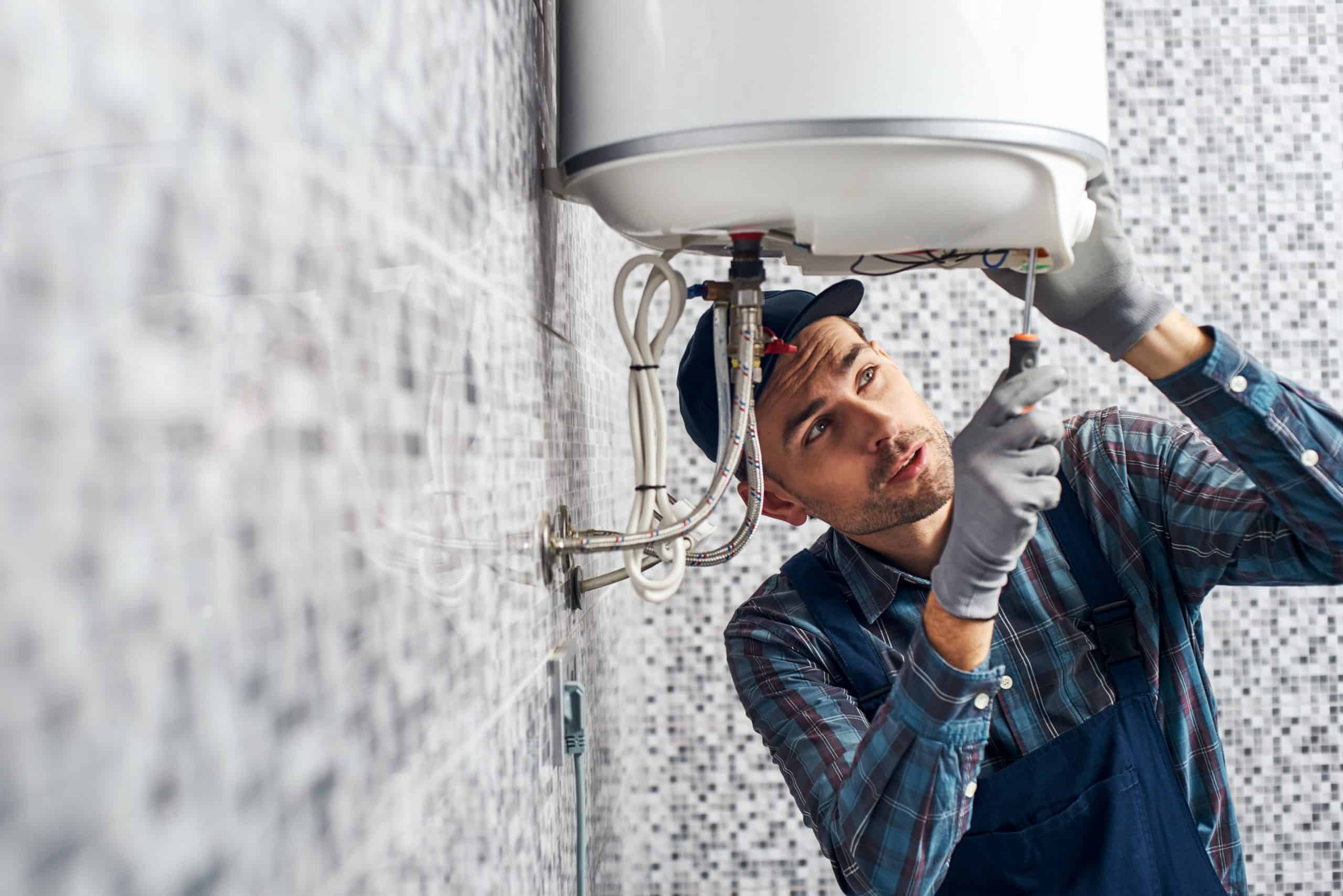 A plumber wearing a cap and overalls works on a water heater mounted on a tiled wall. He uses a screwdriver to adjust parts, focusing intently on the task. The wall is covered with small, square, gray and white mosaic tiles.