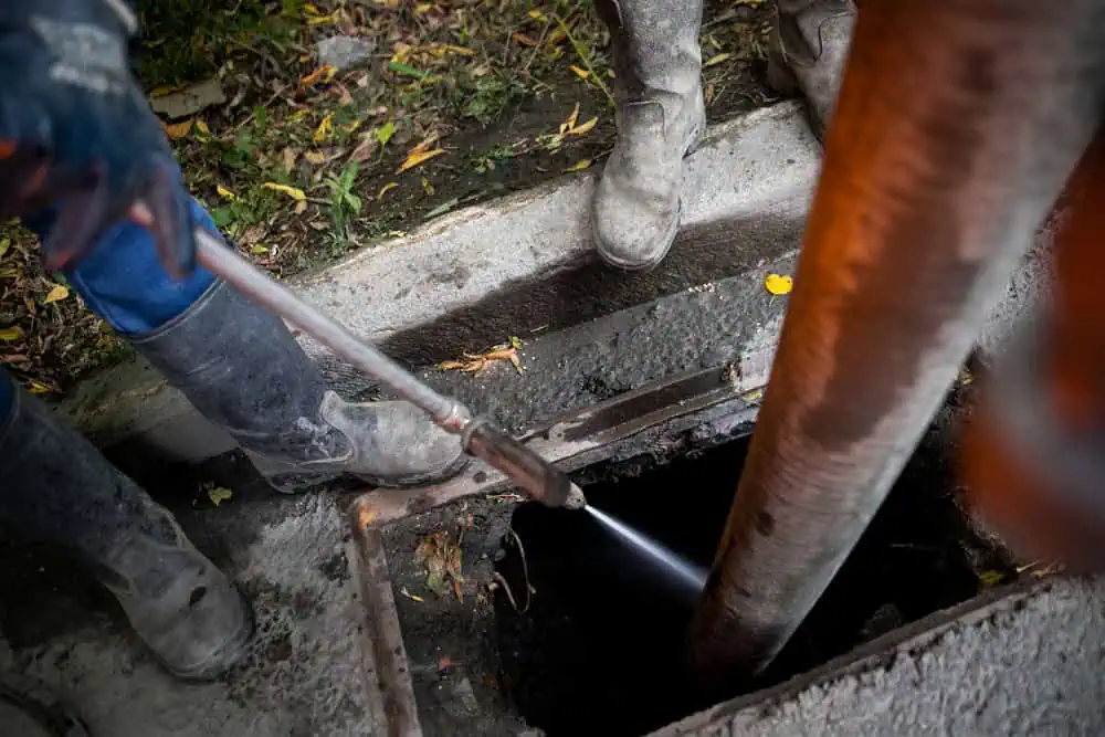 Two workers using a high-pressure hose to clean a street drain. They are wearing protective boots and gloves. The surrounding area has patches of grass and fallen leaves. The hose sprays water into the open drain.