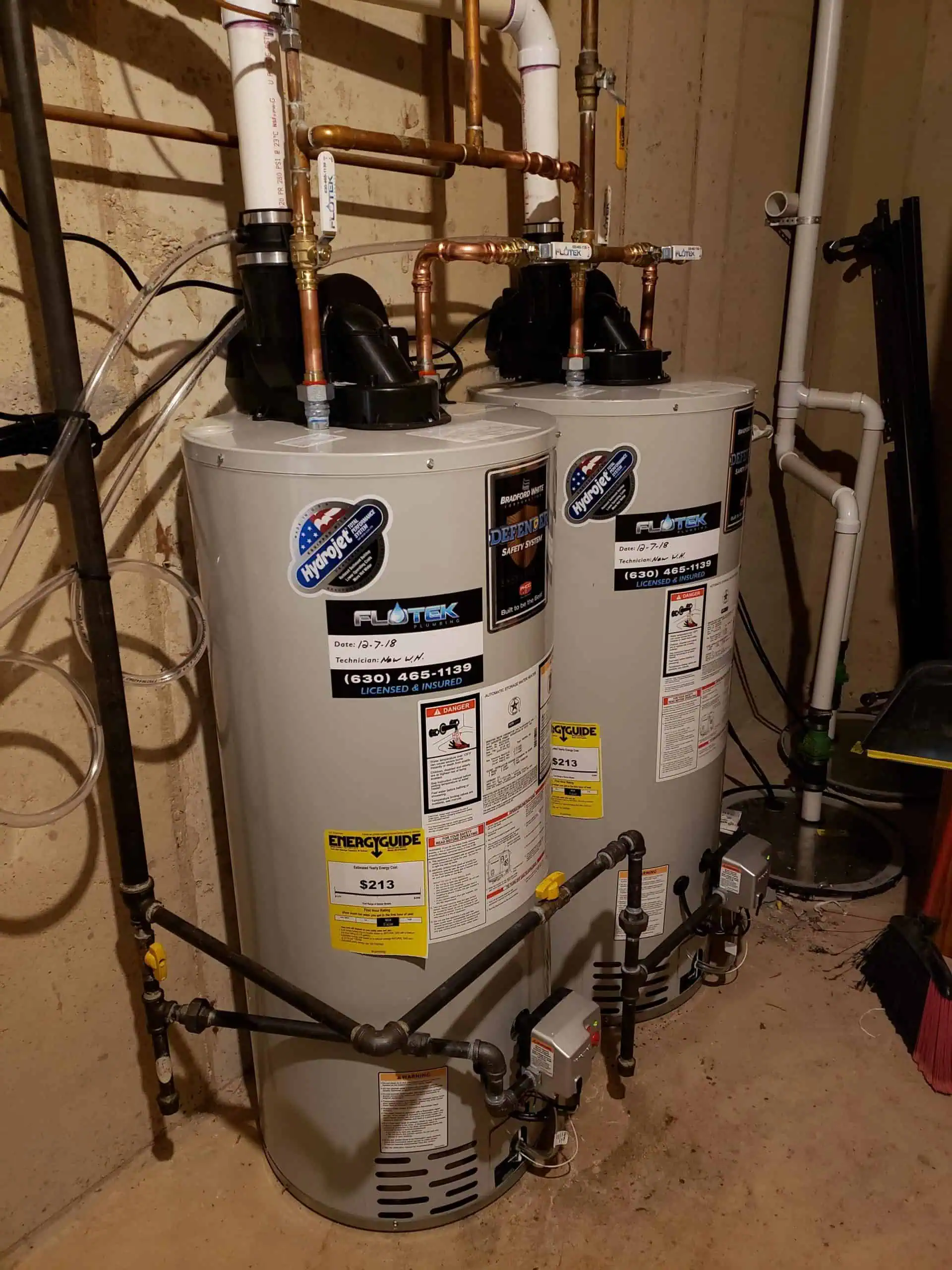 Two large, cylindrical water heaters are installed side by side in a basement. They are connected to various pipes and have labels with brand and efficiency information. The surrounding area has exposed concrete walls and various tools leaning against them.