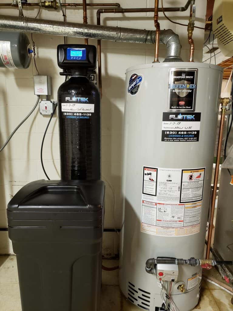 A water softener system with a digital display is next to a large, gray water heater in a basement. Pipes run along the wall, and warning labels are visible on the heater. The floor is concrete.