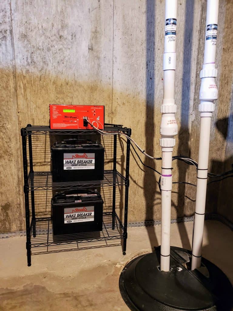 A basement setup with two black batteries labeled "Wake Breaker" on a metal shelf. A red battery maintainer sits on top, with cables connecting to the batteries. Nearby, PVC pipes lead into a circular sump pit on the ground.