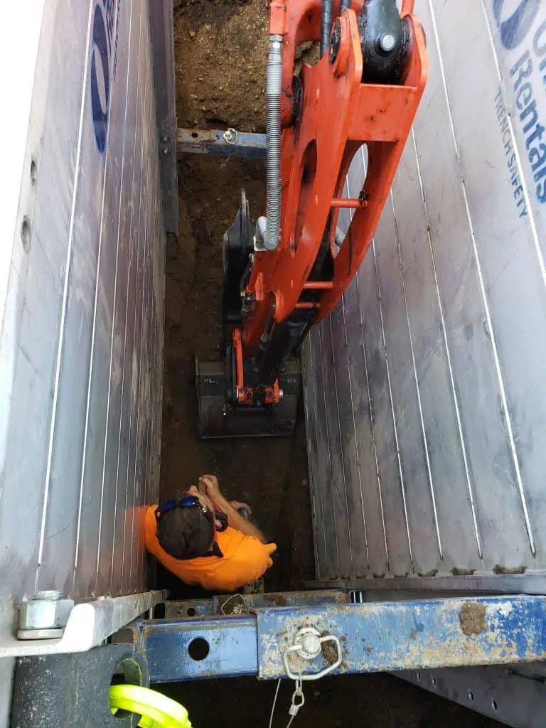 A construction worker in an orange shirt stands in a narrow trench, operating a bright orange excavator. The trench is lined with metal supports for safety. The scene is viewed from above.