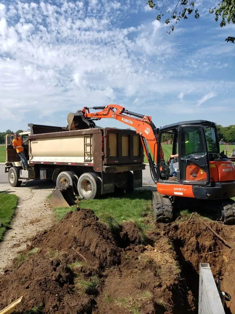 An orange excavator loads dirt into a large dump truck on a construction site. A worker stands on the truck guiding the process. The sky is blue with scattered clouds, and green grass surrounds the area.