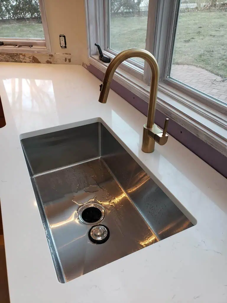A modern kitchen sink with a shiny stainless steel basin and a sleek, curved brass faucet is set in a white countertop. Bathed in natural light from large windows, it offers a lovely view of the grassy yard and walkway. Perfect for any update, consider consulting a reputable plumber near me.
