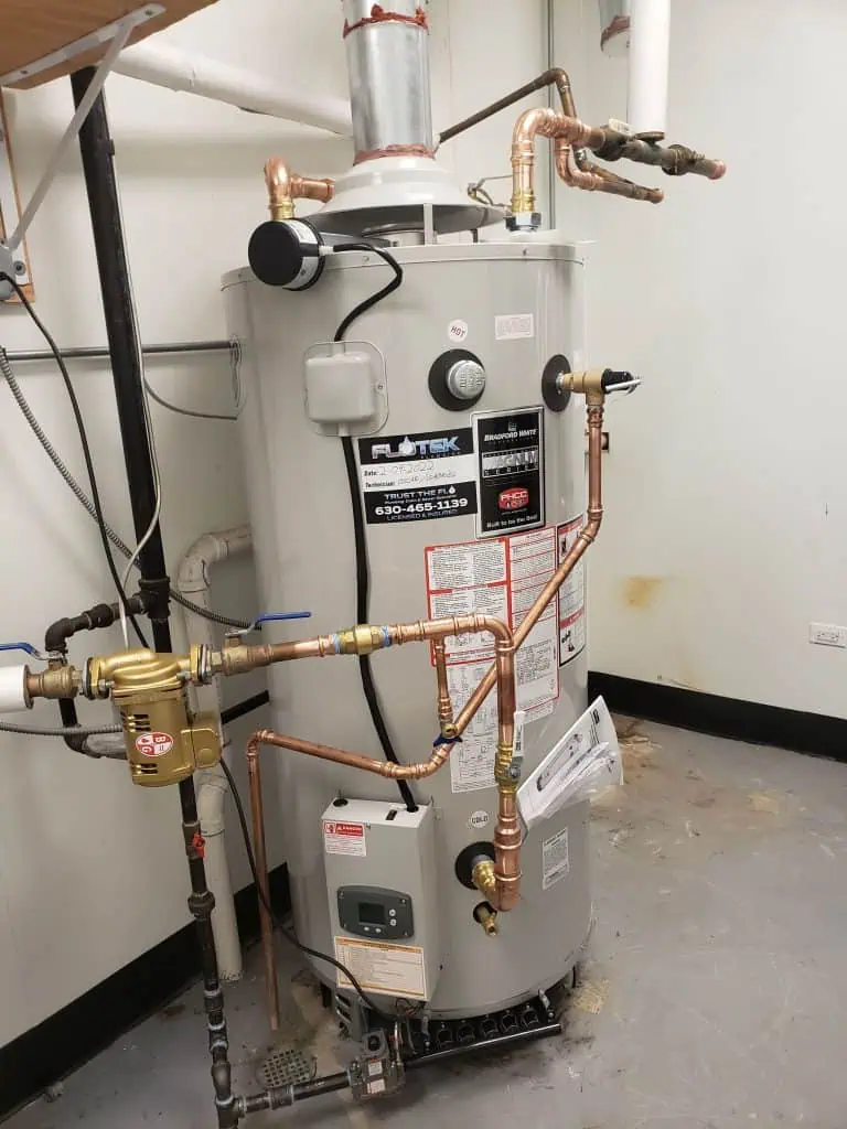 A standing water heater with various copper pipes and valves connected, expertly installed by a plumber near me. The unit is white, sporting multiple labels and warning stickers. It's situated in a utility room with a gray floor and a few wall fixtures.