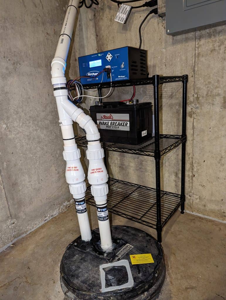 A basement corner with a sump pump system features PVC pipes, a battery backup, and a control unit on a metal shelf. The setup rests against concrete walls and a floor with a circular plastic basin—perfectly installed by your reliable plumber near me.
