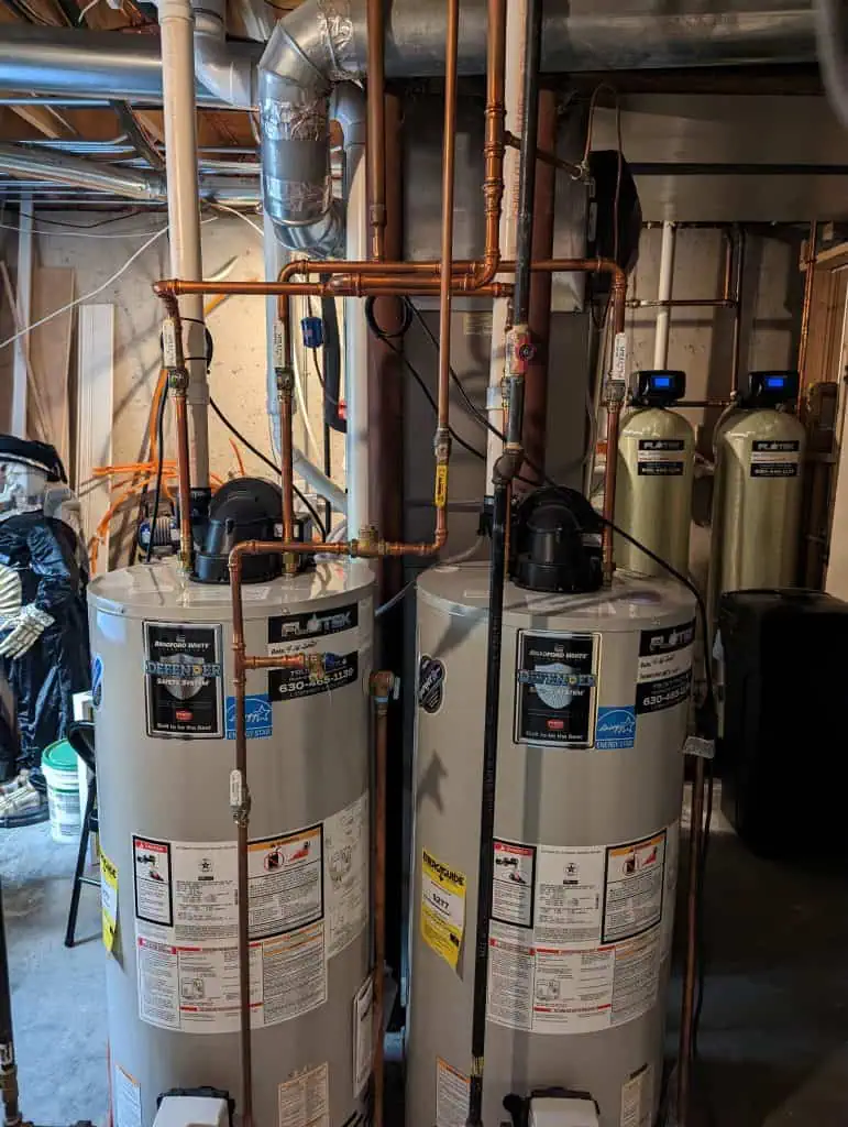 Two water heaters with various labels are installed in a basement. Copper pipes run vertically and horizontally above them. In the background, two water filtration units and miscellaneous basement items are visible.
