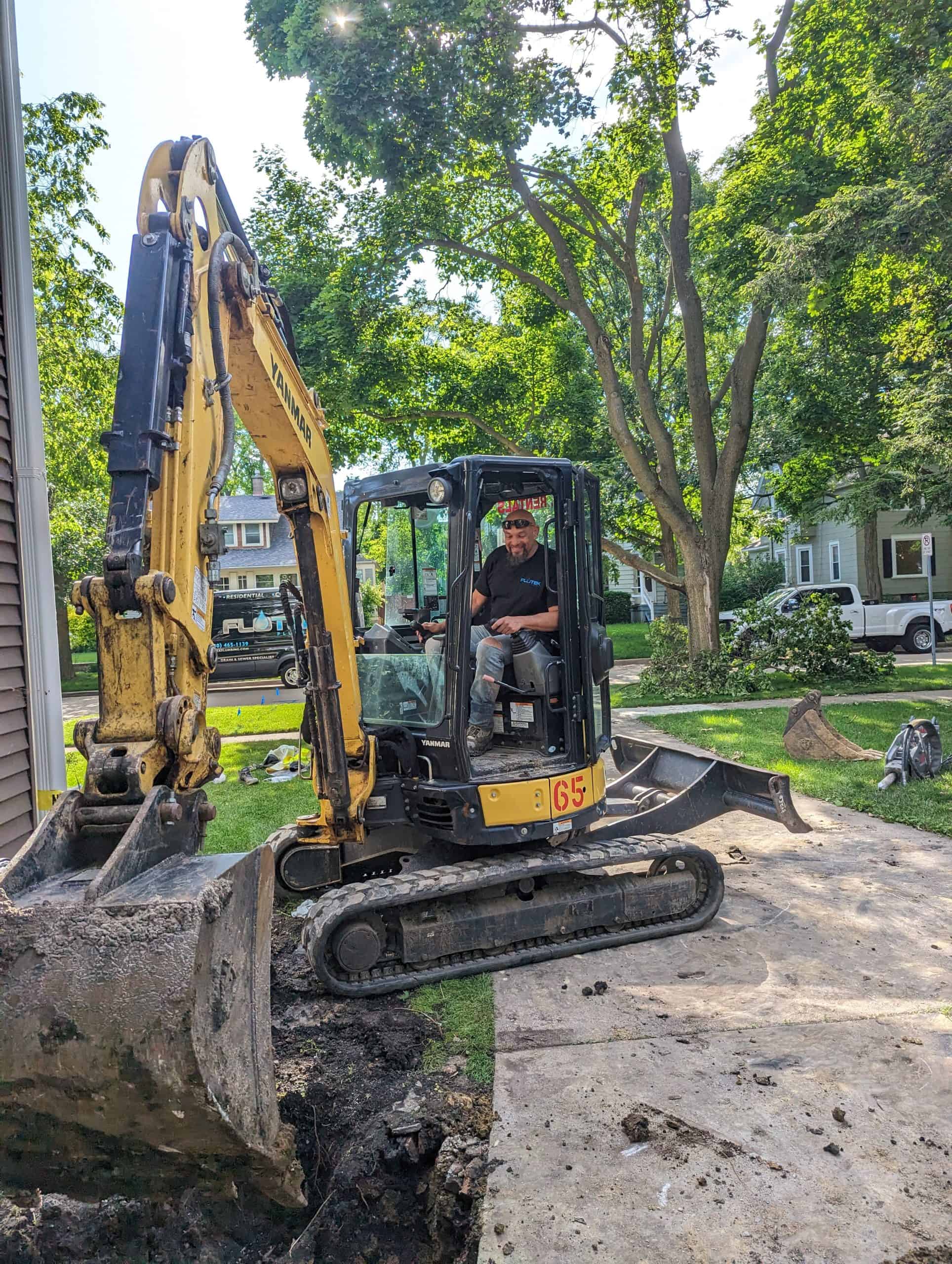 A man operates a small yellow excavator on a residential driveway, digging into the earth. Trees and houses line the street in the background on a sunny day. Equipment and dug-up soil are visible around the site.